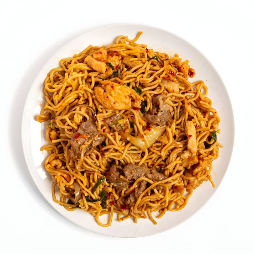 A white plate holds a generous serving of Asian noodles with chicken, beef, and vegetables.